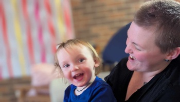 A happy toddler in a blue shirt smiles brightly as a smiling adult with short hair looks on joyfully.