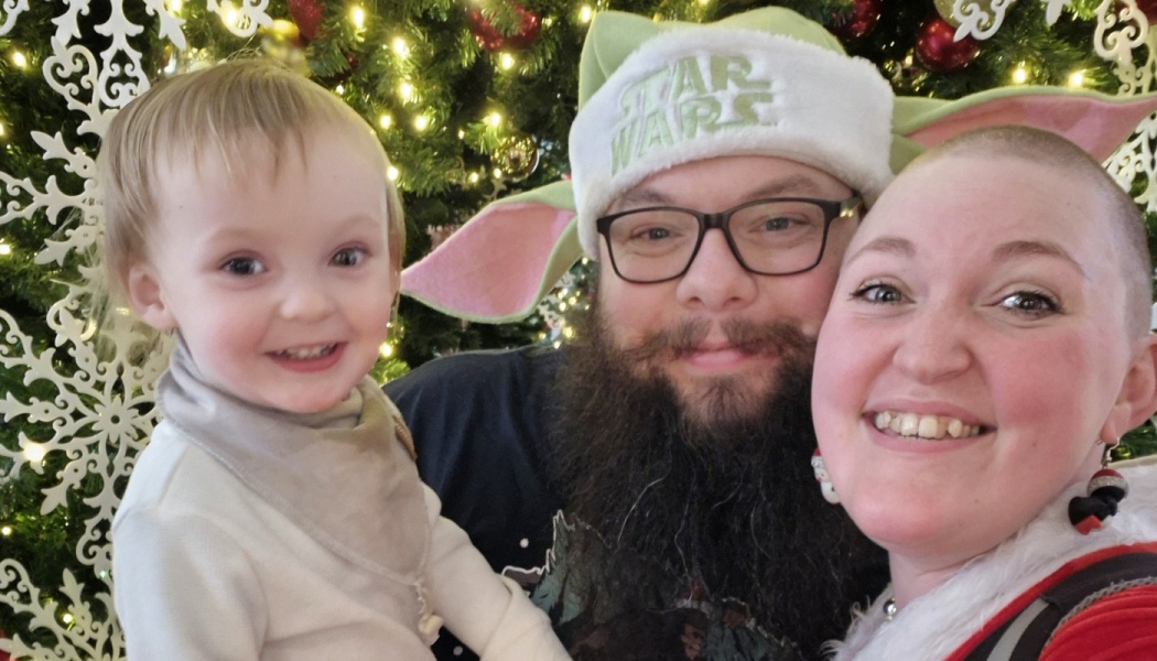 A smiling family of three, including a toddler and a dad in a Yoda Santa hat, posing by a glowing Christmas tree.