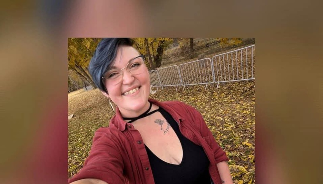 Smiling person with short blue hair and glasses wearing a red shirt outdoors among fallen autumn leaves.
