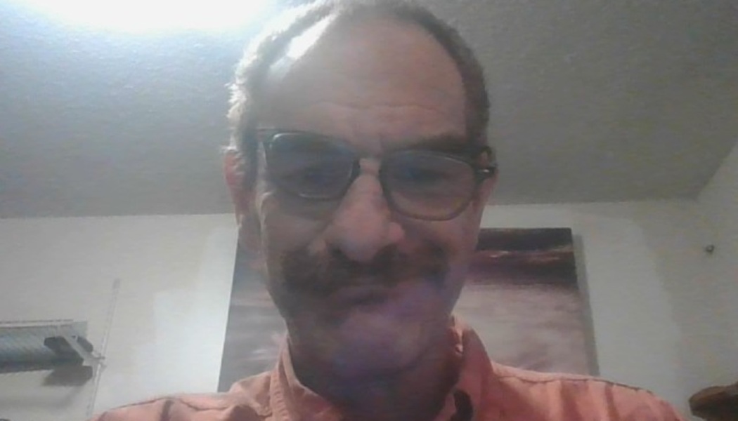 A man with glasses and a mustache wearing a peach button-down shirt looks into the camera.