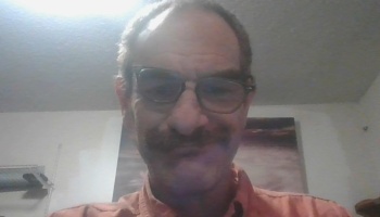 A man with glasses and a mustache wearing a peach button-down shirt looks into the camera.