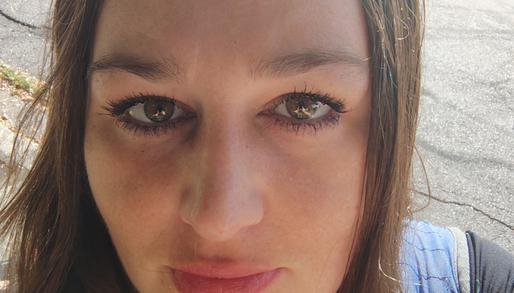 Close-up selfie of a woman with brown eyes and dark hair looking directly at the camera outdoors.