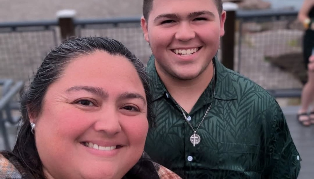 A smiling woman and a young man in a green patterned shirt pose close together for a selfie outdoors.