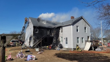 Firefighters respond to a heavily fire-damaged two-story house with smoke rising and toys in the yard.