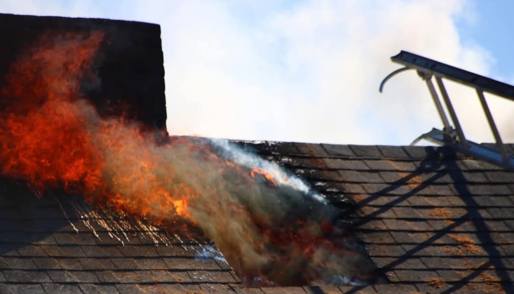 Bright orange flames and thick smoke burst through a hole in a dark shingled roof near a chimney.