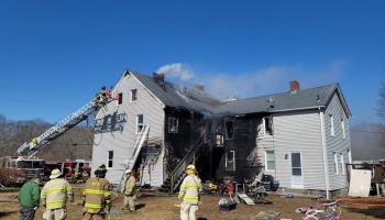 Firefighters and a ladder truck at a heavily charred, fire-damaged two-story house.