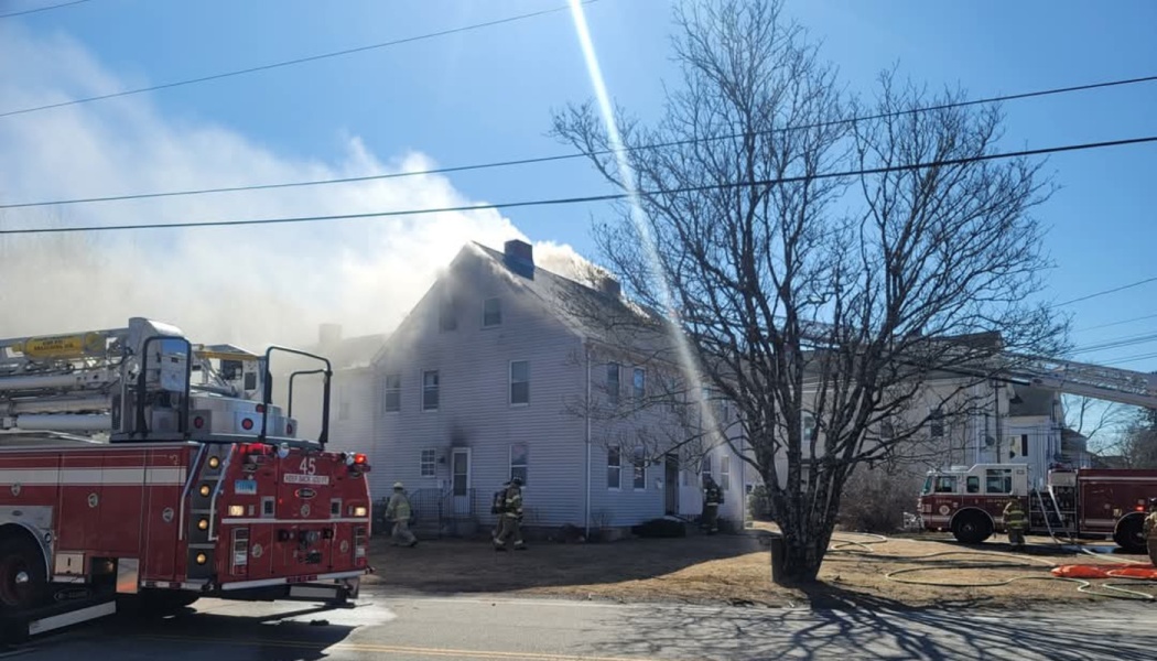 Firefighters and fire trucks respond to a severe house fire with heavy smoke pouring from the roof.