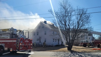 Firefighters and fire trucks respond to a severe house fire with heavy smoke pouring from the roof.