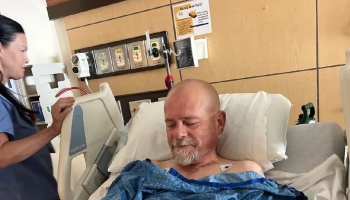 A bald man with a goatee rests in a hospital bed while a nurse stands beside him.