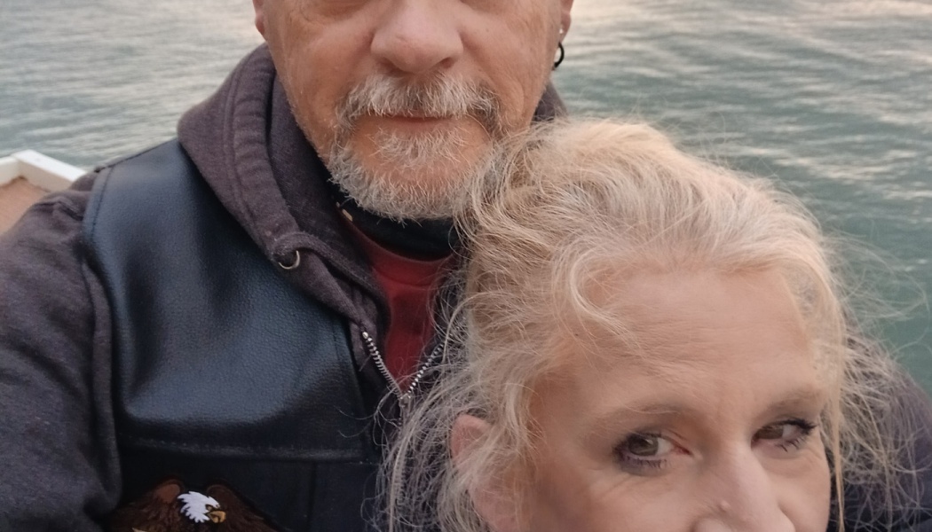 Close-up selfie of a bearded man in a leather vest standing with a blonde woman in front of water.