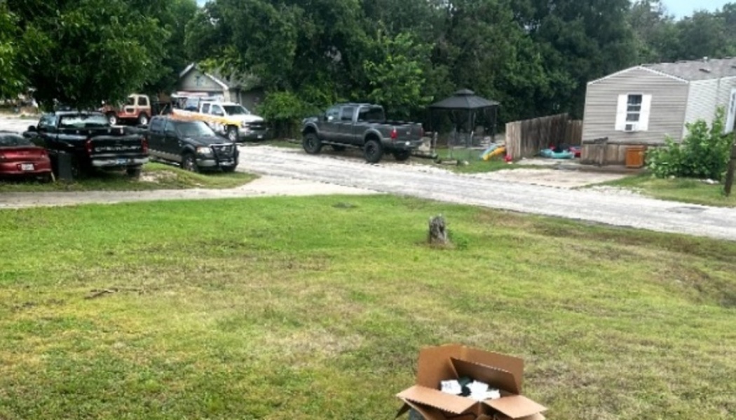 An open cardboard box sits in a grassy yard near a dirt road lined with parked pickup trucks and a mobile home.