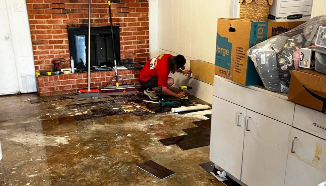 A man uses a power drill to repair water-damaged flooring near a brick fireplace during room renovation.