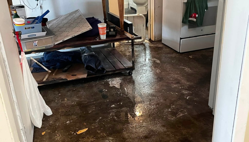 Standing floodwater covers the floor of a home, surrounding furniture, clutter, and kitchen appliances.