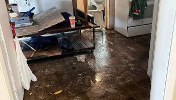 Standing floodwater covers the floor of a home, surrounding furniture, clutter, and kitchen appliances.