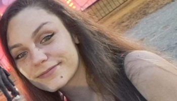Close-up portrait of a young woman with long dark hair and hazel eyes smiling outdoors.