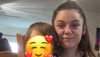 A smiling woman poses next to a young child whose face is covered with a heart emoji.