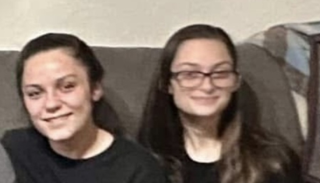 Two young women smiling side-by-side on a couch, one wearing glasses.