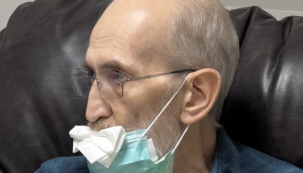 A man with glasses resting in a leather chair, wearing a medical face mask tucked with a tissue.