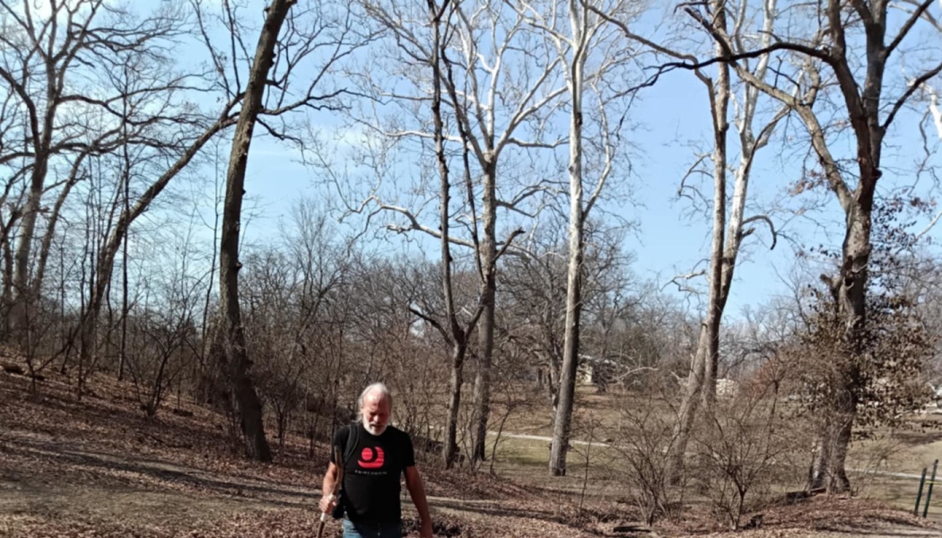 An older man with a white beard hikes through a bare winter forest holding a walking stick.