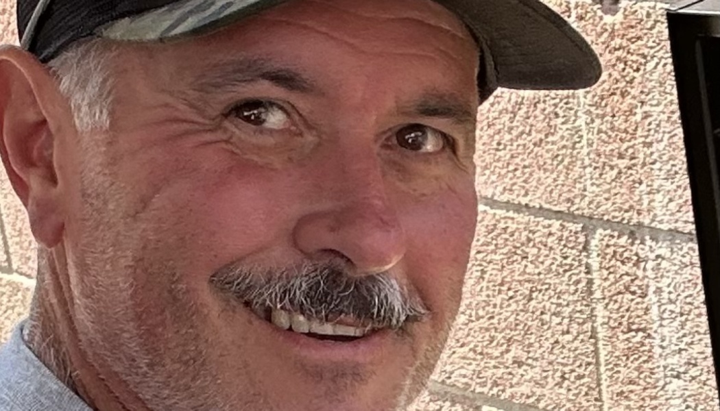 Close-up portrait of a smiling man with a grey mustache wearing a baseball hat.
