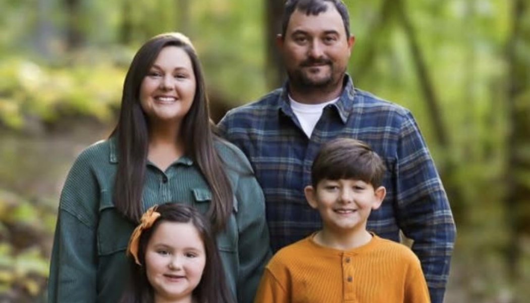 A smiling family of four—a mother, father, son, and daughter—poses together in a wooded outdoor setting.