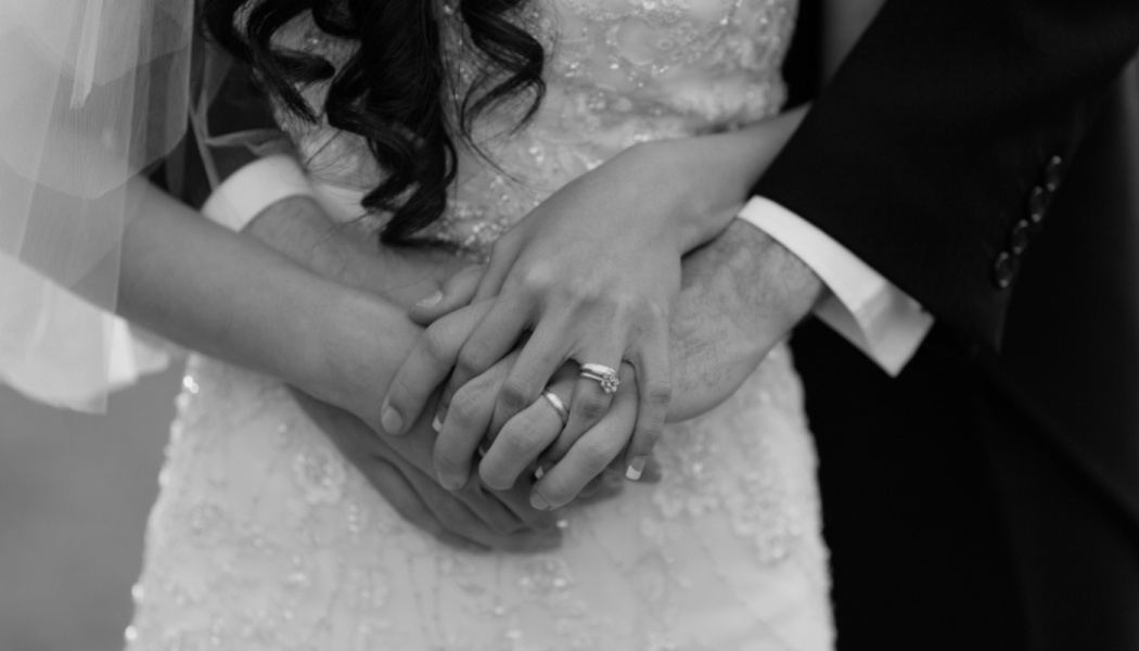 Black and white close-up of a bride and groom holding hands, showcasing their wedding rings against a lace dress.