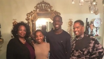 A smiling Black family of four standing together in front of an ornate gold mirror.