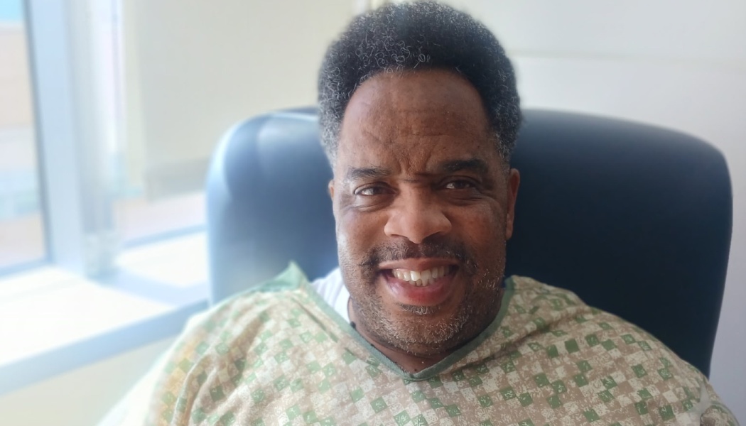 A smiling Black man wearing a patterned hospital gown sits in a chair near a bright window.