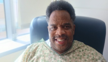 A smiling Black man wearing a patterned hospital gown sits in a chair near a bright window.