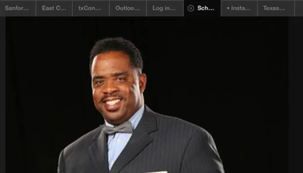 A smiling Black man in a dark suit, light shirt, and grey bowtie against a black background.