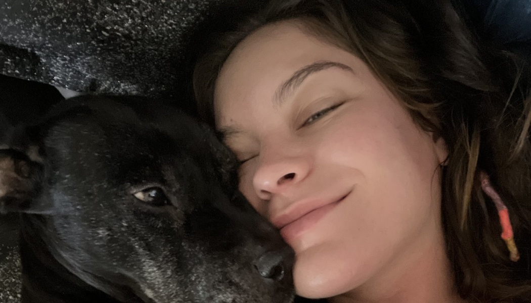 A smiling woman rests her cheek affectionately against her black dog while lying down.