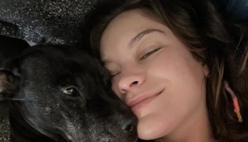 A smiling woman rests her cheek affectionately against her black dog while lying down.
