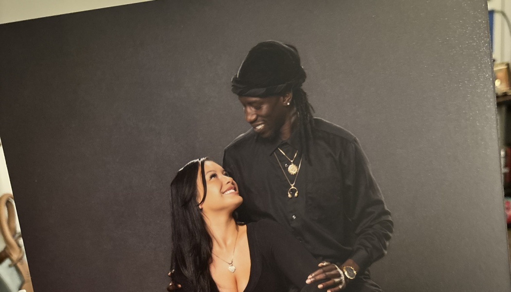 A smiling man and woman dressed in black gaze lovingly at each other against a dark studio backdrop.