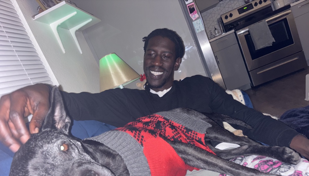 A smiling man relaxes on a couch while petting his black dog wearing a red and grey sweater.