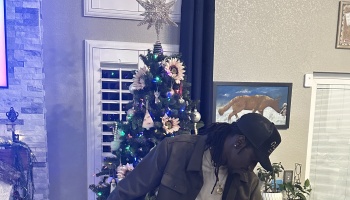 A man with dreadlocks wearing a black cap looks down in front of a decorated Christmas tree topped with a star.