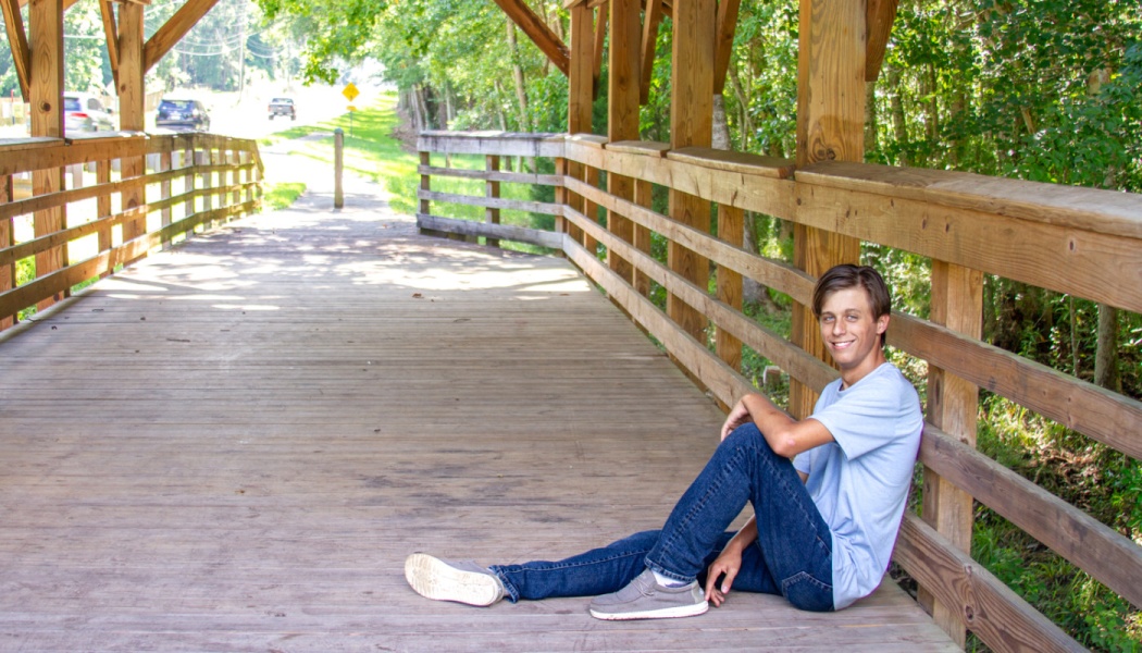 A smiling young man in a blue t-shirt and jeans sitting on a wooden covered bridge.