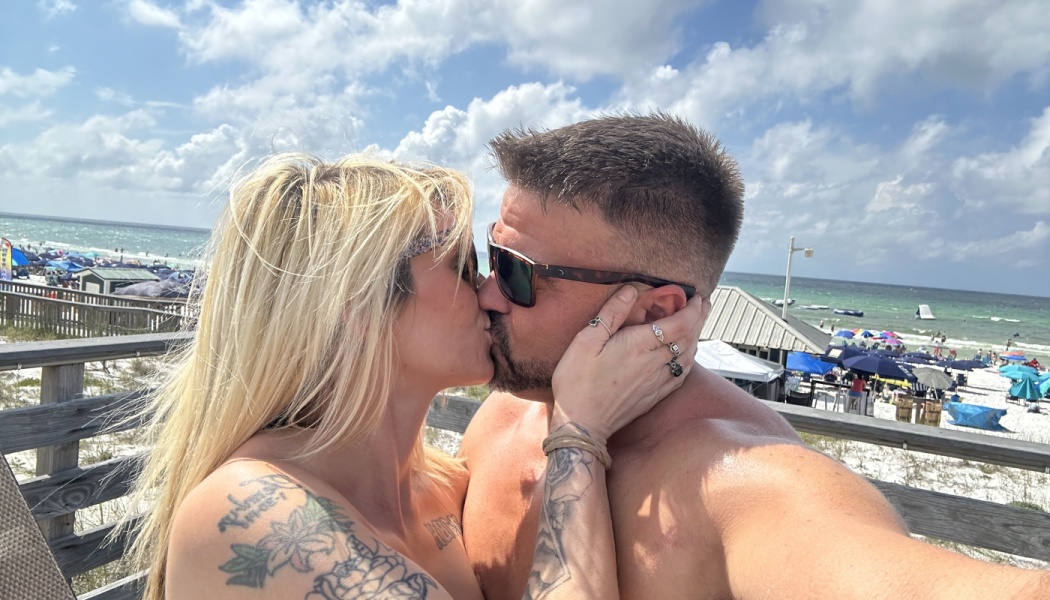 A tattooed couple kisses on a sunny boardwalk with a crowded beach and ocean in the background.