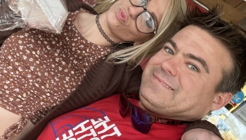 A smiling man with dark hair and a blonde woman wearing glasses making a playful face in a close-up selfie.
