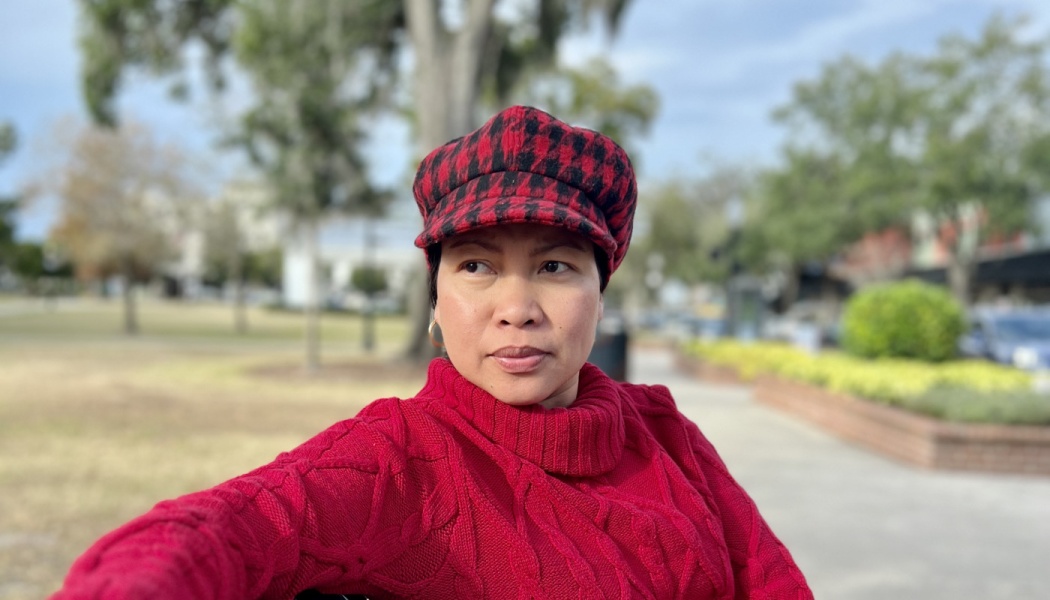 Woman wearing a red cable-knit sweater and red-and-black plaid cap looking thoughtfully off-camera outdoors.