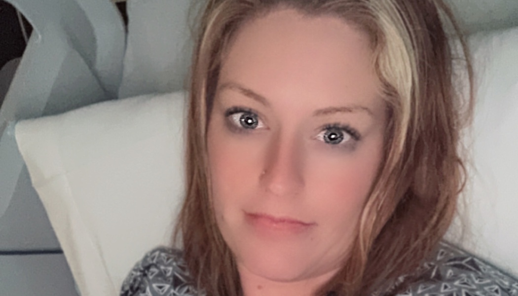 A woman with blonde-highlighted hair lying in a hospital bed, looking directly at the camera.