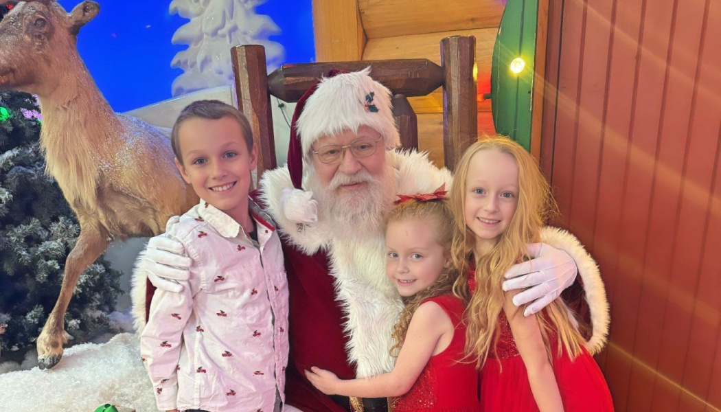 Three smiling children pose happily with Santa Claus in a festive holiday display.