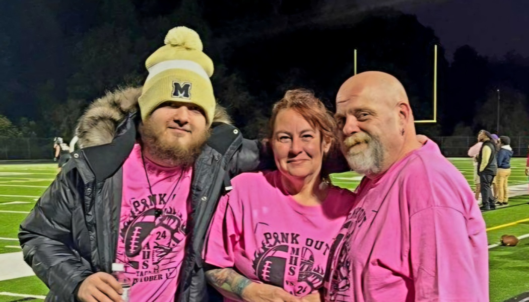 Three people smiling together on a football field at night, wearing matching pink "Pink Out" shirts.