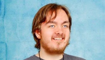 Smiling young man with light brown hair, blue eyes, and facial hair against a textured blue background.