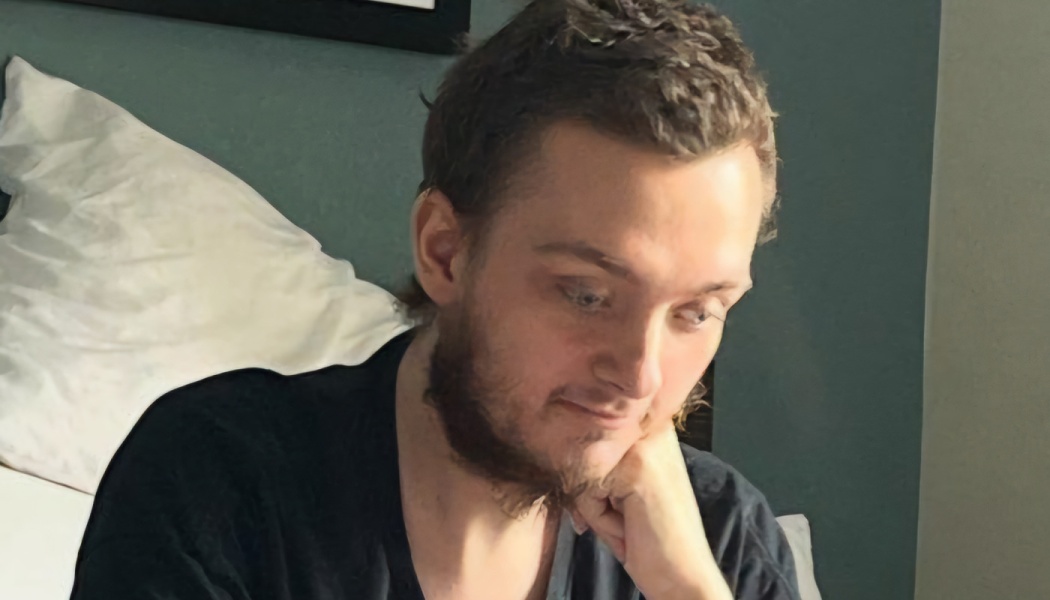 A young man with short brown hair and a beard resting his chin on his hand with a pensive expression.