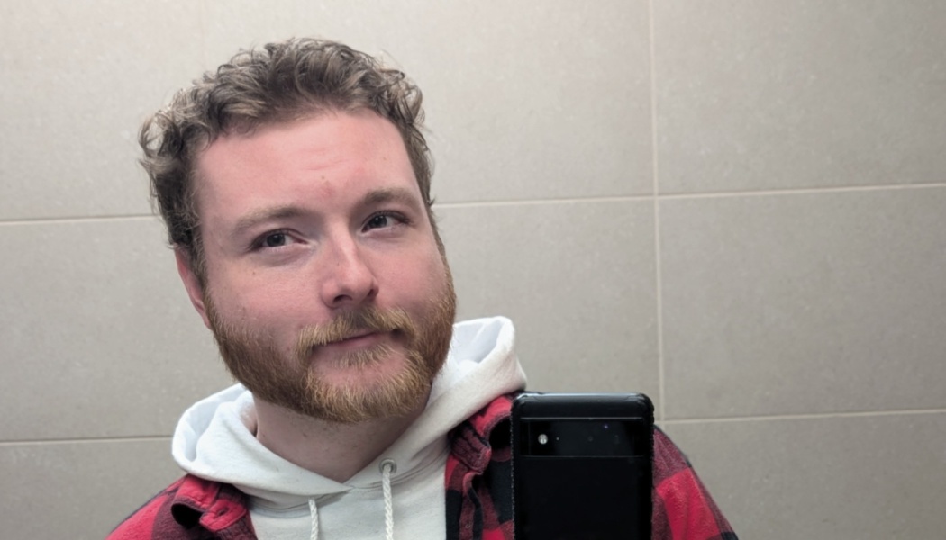 Young man with curly hair and a beard taking a mirror selfie wearing a white hoodie and red plaid shirt.