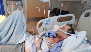A tattooed man rests in a hospital bed with a breathing tube, chest monitors, and medical equipment attached.