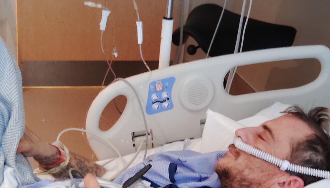 A man lying in a hospital bed with eyes closed, wearing a breathing hose and attached to IV lines.