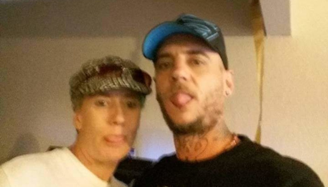 Two men wearing hats playfully stick their tongues out for a selfie indoors.