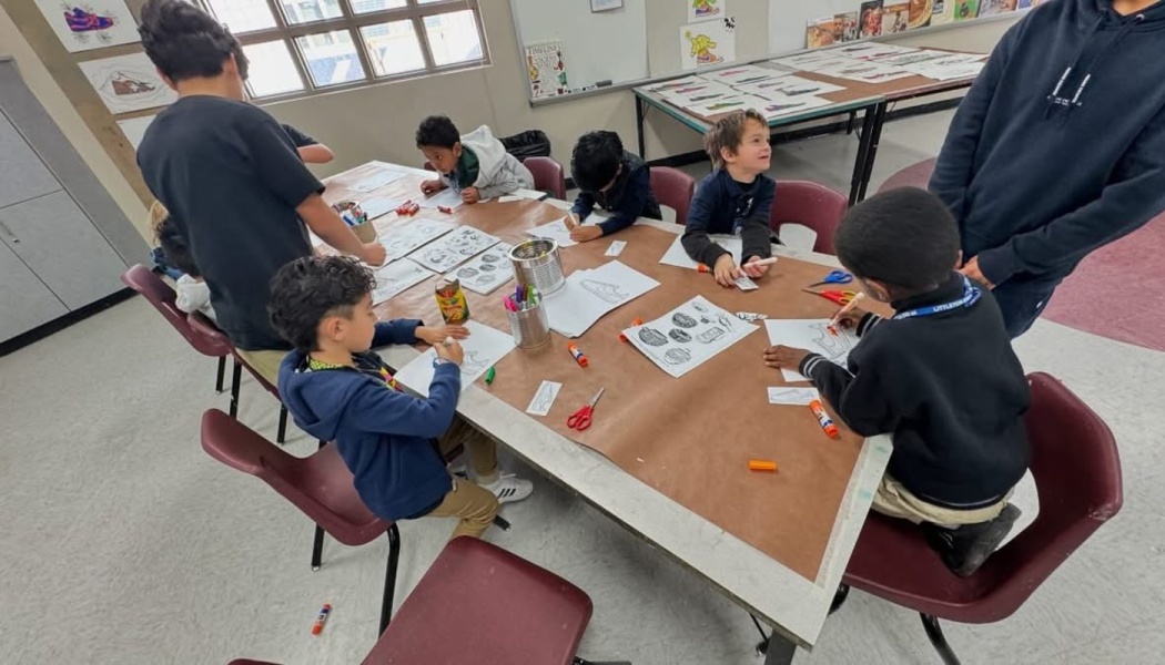 Elementary students gathered around a large table doing art projects with paper, markers, and glue.