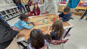 Elementary students collaborate on a large colorful shoe drawing using markers at a classroom table.